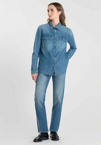 Denim shirt in light blue with button closure, two chest pockets, and long sleeves. Paired with straight-leg light denim jeans.