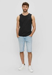 Schwarzes Baumwoll-Tanktop, hellblaue Jeansshorts und weiße Sneakers. Das Model steht mit Händen in den Taschen und präsentiert die lässige Sommerbekleidung.