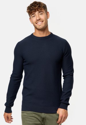 Suéter de punto azul marino con cuello redondo, patrón texturizado, puños y dobladillo de canalé. Combinado con pantalones verde claro.