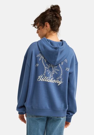 Femme aux cheveux bouclés attachés porte un sweat à capuche bleu avec un motif de palmier et le texte "Billabong 1973" dans le dos, assorti à un jean bleu.