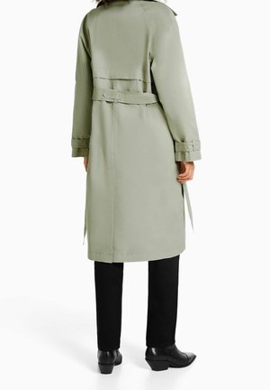 Person iført en lang salviegrøn trenchcoat med bælte, sorte bukser og sorte hælede sko, stående mod en hvid baggrund.