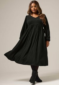 Vestido largo negro de manga larga con escote en V, tejido texturizado, cintura fruncida y falda fluida. Combinado con botas negras hasta el tobillo.