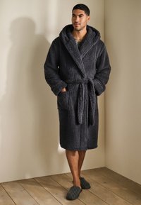 Donkergrijze, zachte badjas met capuchon, geknoopt aan de taille, met zijzakken. Draagt over lichtgekleurde kleding op een houten vloer.