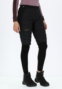 Zwarte gewatteerde shorts met daaronder zwarte leggings, voorzien van een goudkleurig logo op de dij, gedragen met zwarte wandelschoenen.