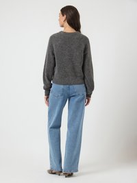 Mulher virada de costas, vestindo um suéter cinza escuro felpudo, calças jeans retas azuis claras e sapatos de salto bége contra um fundo simples.
