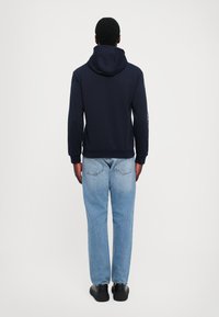 Felpa con cappuccio blu navy dal taglio rilassato, con polsini a costine e tasca a marsupio, abbinata a jeans azzurri a gamba dritta e scarpe nere.