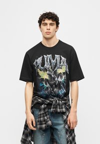 Νεαρός άνδρας με σγουρά μαλλιά, φοράει μαύρο υπερμεγέθες t-shirt Puma με γραφικό σχέδιο πάνθηρα και καρό πουκάμισο δεμένο γύρω από τη μέση.