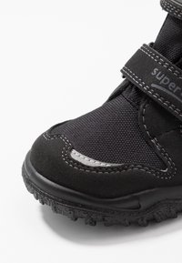 Botín negro impermeable con parte superior texturizada, strap de Velcro, puntera reforzada y suela de goma resistente con bandas de tracción.
