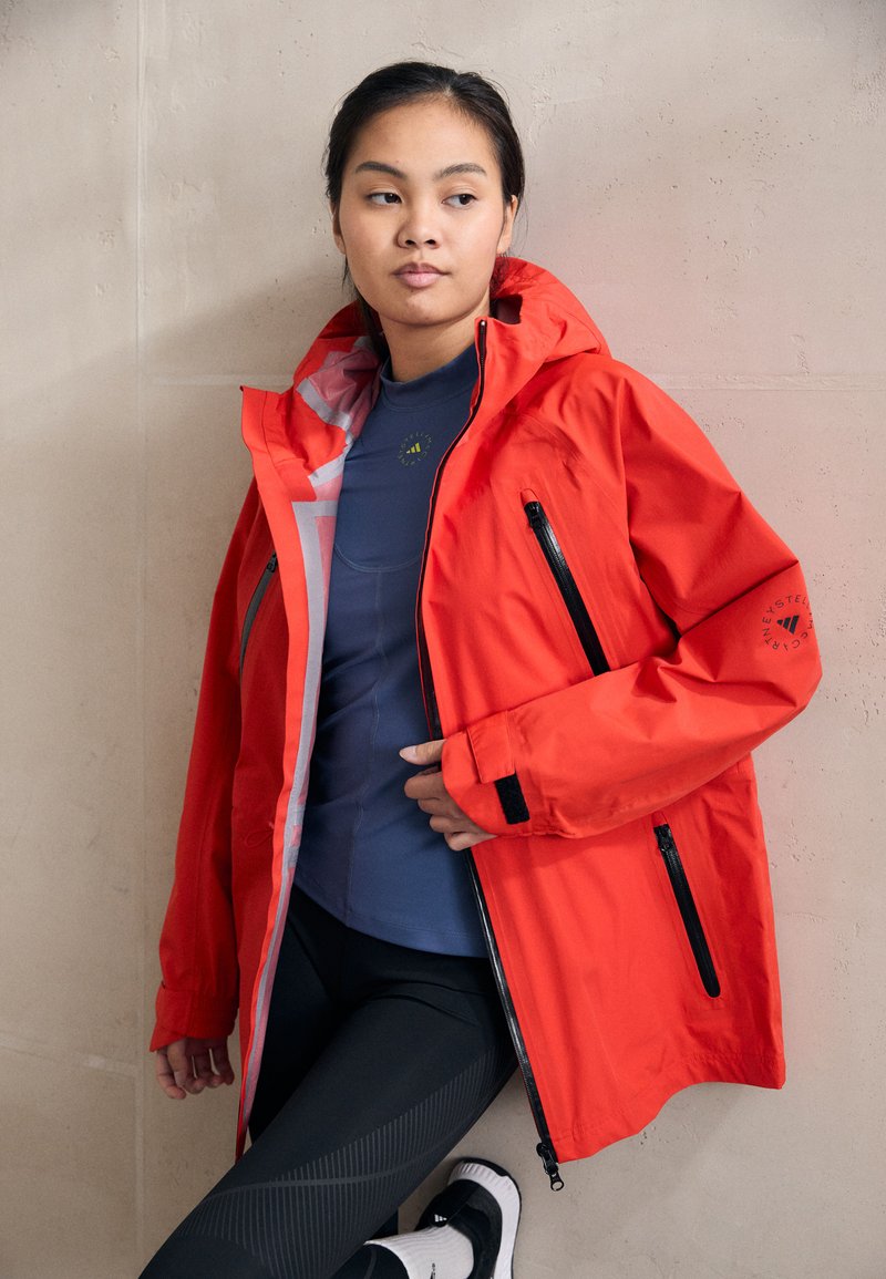 Bright red waterproof jacket with a hood, contrasting inner lining, zippered pockets, and adjustable cuffs worn over a fitted blue top and leggings.