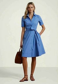 Γυναίκα που φοράει ένα μπλε φόρεμα με κουμπιά μέχρι το γόνατο και κοντά μανίκια, με δεμένο στη μέση, κρατάει μια καφέ τσάντα χειρός και φοράει καφέ σανδάλια με λουράκια.