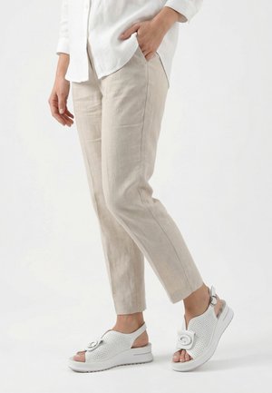 Person trägt beige verkürzte Hose und weiße perforierte Slingback-Sandalen mit Schnallen, steht vor einem schlichten weißen Hintergrund.