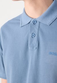 Jasnoniebieska koszulka polo wykonana z fakturowanej tkaniny. Posiada kołnierzyk, zapięcie na dwa guziki oraz subtelne logo w tym samym kolorze na piersi.