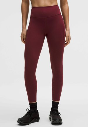 Femeie purtând colanți burgundy cu talie înaltă, pantofi sport negri și șosete, stând într-un fundal deschis simplu.