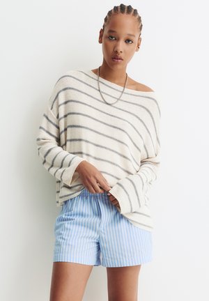 Frau mit geflochtenem Haar, die einen lockeren weißen Pullover mit grauen Streifen und hellblauen gestreiften Shorts trägt, steht vor einem weißen Hintergrund.