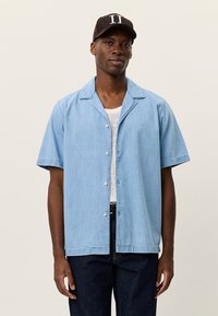 Ανοιχτό μπλε πουκάμισο με κοντό μανίκι και γιακά, με διακριτική υφή και τρία κουμπιά. Φορεμένο πάνω από άσπρη φανέλα και σε συνδυασμό με σκούρα τζιν.