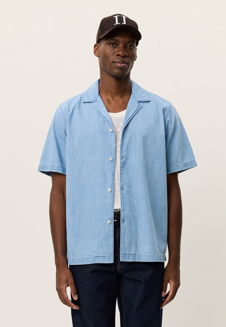 Ανοιχτό μπλε πουκάμισο με κοντό μανίκι και γιακά, με διακριτική υφή και τρία κουμπιά. Φορεμένο πάνω από άσπρη φανέλα και σε συνδυασμό με σκούρα τζιν.