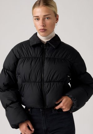 Jeune femme aux cheveux blonds portant une doudoune noire et un col roulé blanc sur un fond uni.