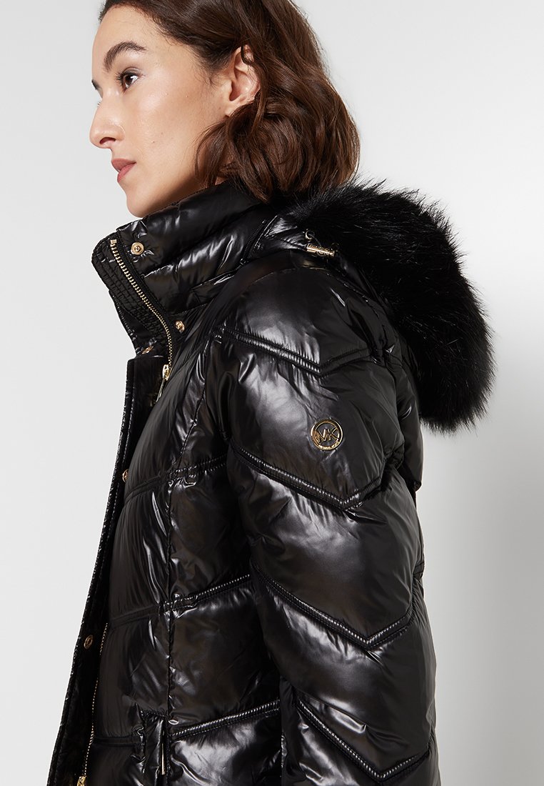 Giacca a vento nera con texture lucida, cuciture a zig-zag, cappuccio foderato in pelliccia e dettagli in hardware dorati.
