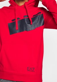 Czerwona bluza z kapturem z czarnym, gumowanym logo na klatce piersiowej, sznurkami i przednią kieszenią kangurka. Miękki materiał, luźny krój.