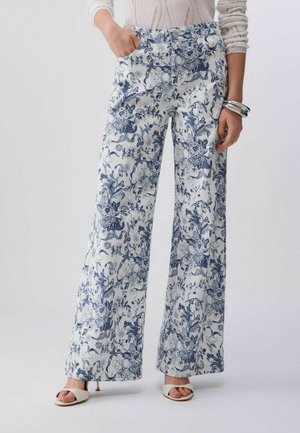 Kobieta w białych spodniach o szerokich nogawkach z niebieskim kwiatowym wzorem, białych sandałach, białym dzianinowym topie i srebrnych bransoletkach na prawej ręce.