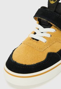 Suede sneaker featuring a beige upper, black accents, white laces, and a cushioned sole with an orange stripe. Velcro strap for secure fit.