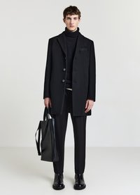 Schwarzer Wollmantel mit Knopfverschluss, getragen über einem schwarzen Rollkragenpullover. Schwarze, eng anliegende Hose und eine kantige Leder-Handtasche.