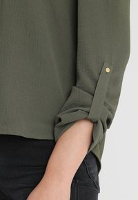 Camicia verde oliva con texture e maniche arrotolate, dotata di chiusura a linguetta e dettagli con bottoni dorati. Abbinata a pantaloni neri.