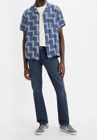 Camicia a maniche corte a quadri blu con dettagli bianchi, indossata sopra una t-shirt bianca e jeans di denim scuro, completata da sneaker grigie.