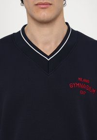 Sweatshirt azul-marinho com decote em V e acabamento branco em contraste, apresentando texto bordado em vermelho a dizer "MILANO GYMNASIUM EA7." Textura suave, design casual.