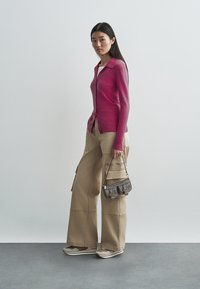 Casaco cardigan rosa de manga comprida, calças cargo bege e mala cinzenta com detalhes dourados. A modelo usa ténis brancos com detalhes bege. Design simples, textura suave.