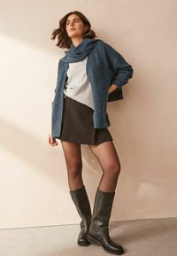 Cardigan bleu tricoté par-dessus un haut gris, une jupe noire et des bottes noires à hauteur de genou avec accent de boucle. Textures lisses, design superposé.