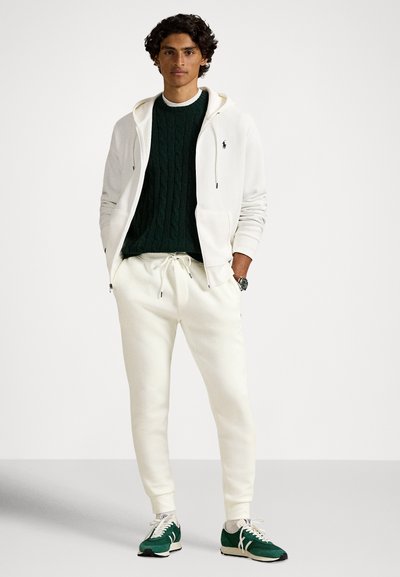 Λευκή ζακέτα με φερμουάρ πάνω από πράσινο σκούρο πουλόβερ πλεχτό με σχέδιο κοτσίδας, συνδυασμένη με λευκά αθλητικά παντελόνια και πράσινα αθλητικά παπούτσια. Ξεχωριστές υφές και άνετη, casual εφαρμογή.