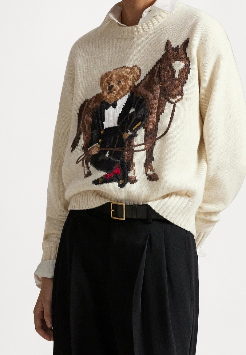 Pull avec design tricoté d'un ours en smoking tenant des rênes, debout à côté d'un cheval marron, porté sur une chemise blanche.