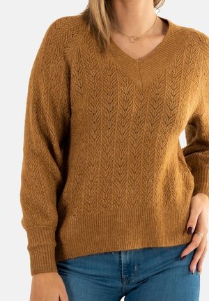 Femme portant un pull en maille marron texturé à col en V et un jean bleu, la main gauche reposant sur la hanche.