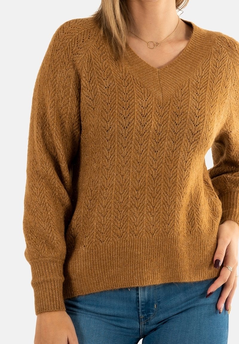 Femme portant un pull en maille marron texturé à col en V et un jean bleu, la main gauche reposant sur la hanche.