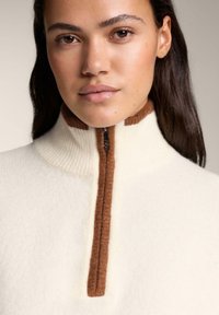 Bijeli flis pulover s visokim ovratnikom i polu-rajfešlusom. Smeđi rebrasti detalji na ovratniku i rajfešlusu. Meka tekstura, minimalistički dizajn.