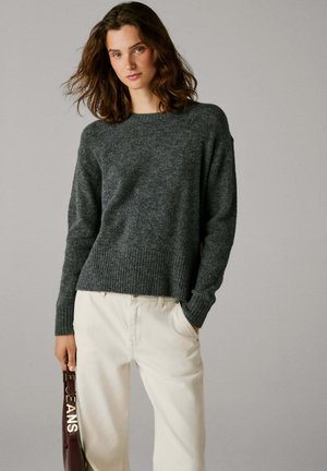 Grijs gebreid sweater met een geribbeld zoom en manchetten, gecombineerd met lichtgekleurde broek. De textuur lijkt zacht en knus.