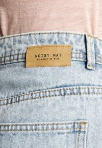 Jeans de denim azul claro com um rótulo de couro a dizer "NOISY MAY" e "SEJA BARULHENTO SEJA LEGAL". Costura visível e detalhes de textura no tecido.