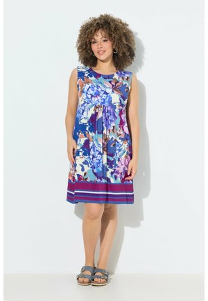Kvinde med krøllet hår iført en ærmeløs blomsterkjole med blå, lilla og pink nuancer, stående mod en ensfarvet hvid baggrund.