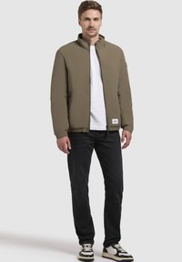 Oliven grøn polstret jakke med ståkrave, lynlås foran, sidelommer og hvidt logo-patch, sat sammen med sorte jeans og sort-hvide sneakers.