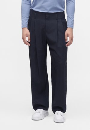 Homme portant un pantalon noir ample, une chemise bleu clair à manches longues et des baskets blanches, debout devant un fond uni.