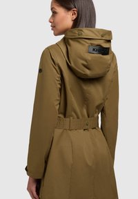 Maslinasto zeleni parka s kapuljačom svezanom u struku, remenom u struku i teksturiranim materijalom. Ima logotip na leđima i dugmad na preklapanje.