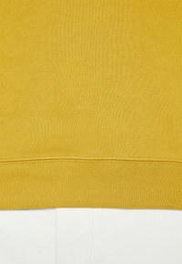 Żółty sweter wykonany z prążkowanej tkaniny z prostym dołem. Dolna krawędź ma staranne wykończenie, kontrastujące z teksturowanym białym podłożem poniżej.