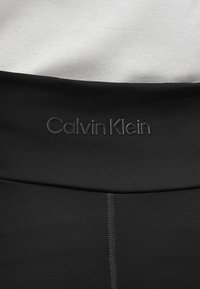 Talie din material negru elastic cu logo „Calvin Klein” embosat, asortată cu un top alb neted și texturat. Linii curate și design minimalist.