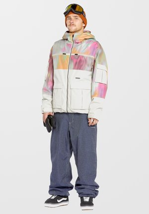 Chaqueta de nieve con patrón de mármol en tonos pastel, sección inferior blanca, múltiples bolsillos, combinada con pantalones de denim holgados y zapatillas negras.