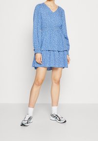Robe bleue à pois blancs, manches longues, col en V, taille froncée, jupe à volants. Portée avec des chaussettes blanches et des baskets multicolores.