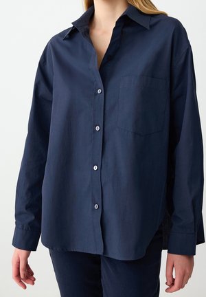 Femme portant une chemise ample bleu foncé avec boutons, poche poitrine et manches longues, assortie à un pantalon bleu foncé.