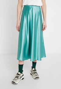 Tyrkysová saténová midi sukně s hladkou texturou, vybavená elastickým pasem a splývavou siluetou, stylizovaná s barevnými ponožkami a teniskami.