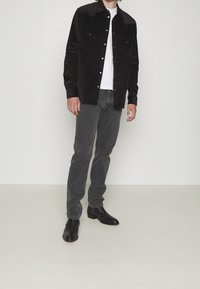 Camicia nera in corduroy con bottoni e tasche frontali, abbinata a jeans grigi e stivali neri in pelle alla caviglia. In primo piano su uno sfondo semplice.