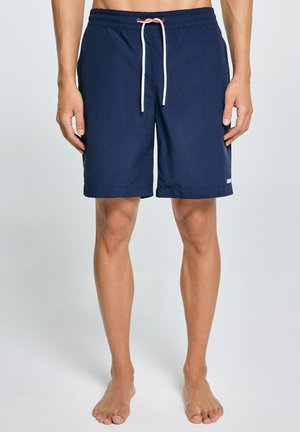 Mężczyzna ubrany w granatowe kąpielówki z białym sznurkiem, stojący boso na tle jednolitego jasnego tła.